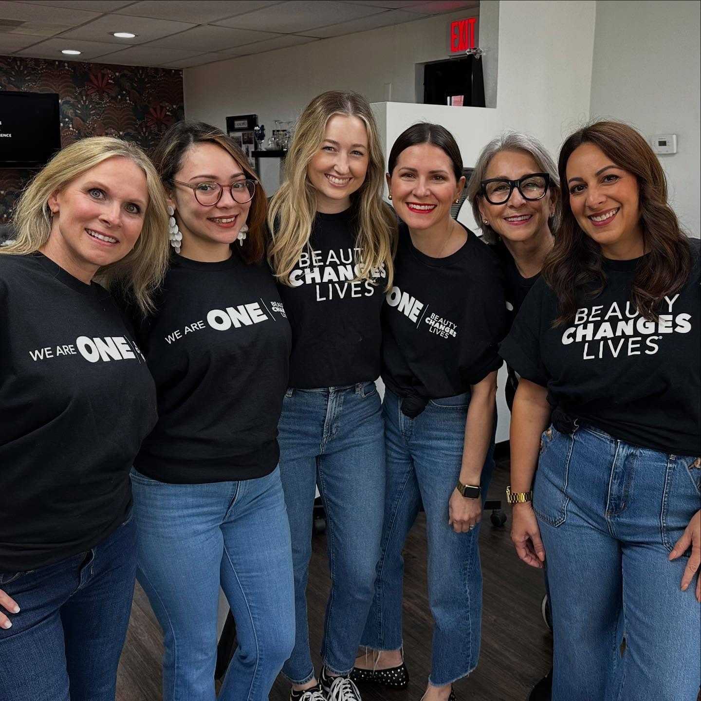 Group of smiling women in matching shirts and jeans, gathered in an indoor setting.