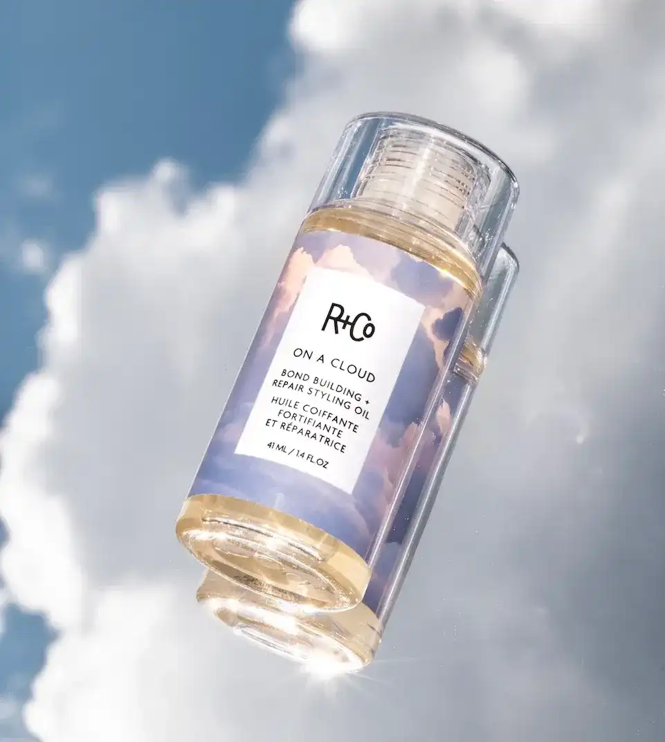 R+Co On a Cloud hair oil bottle with sky and clouds backdrop.